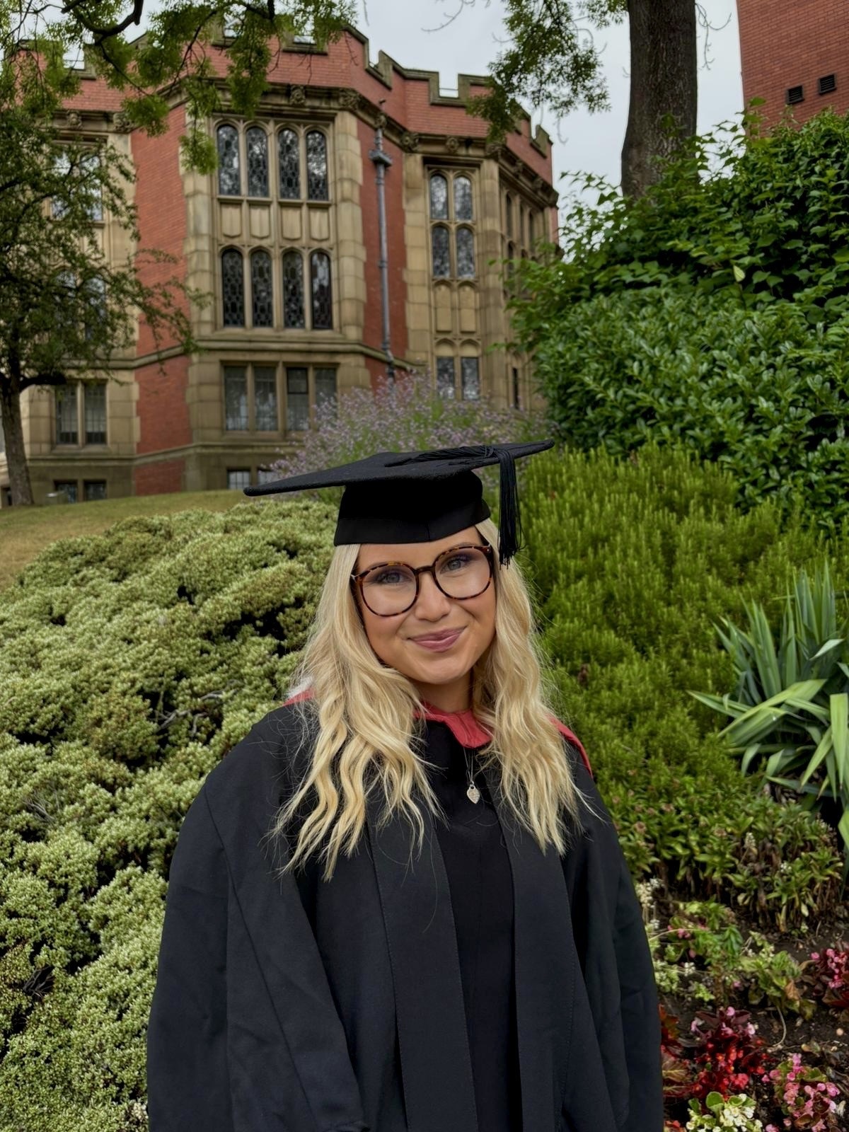 Alex wearing her graduation cap and gown stood outside a university building surrounded by trees and green plants. 
